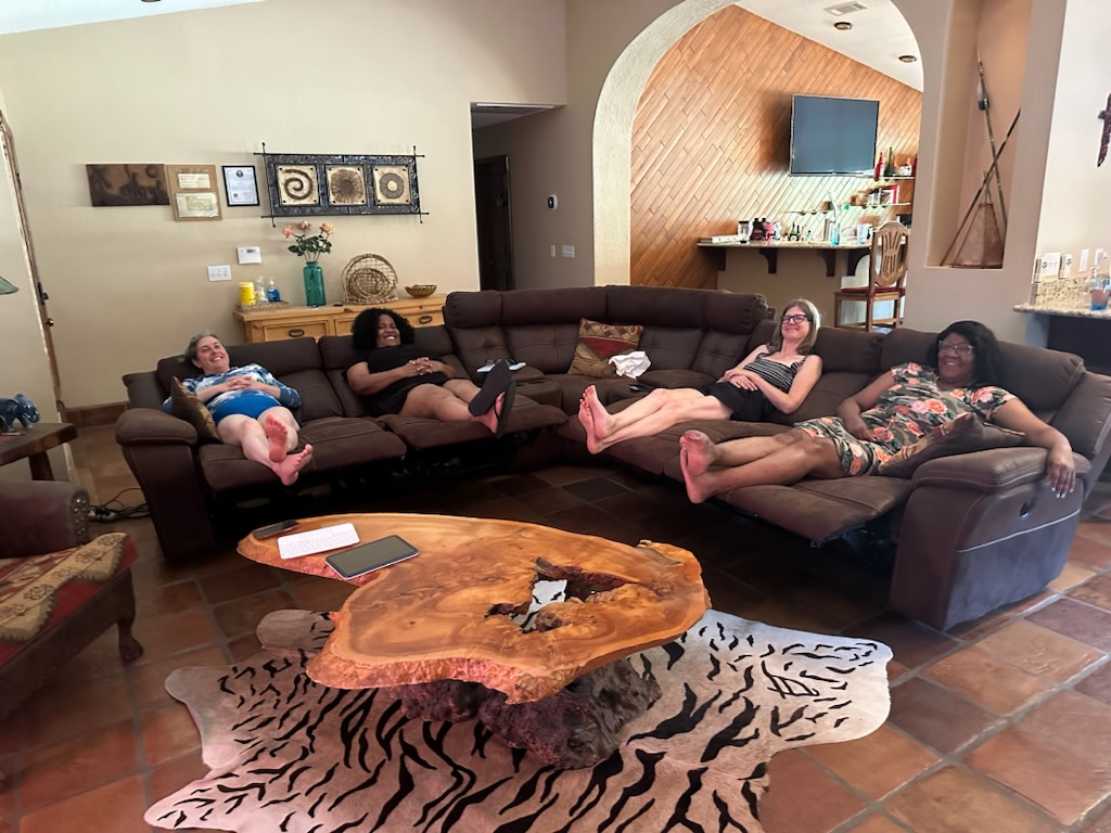 Four women recline on the reclining seats of a C-shaped couch, smiling. In the foreground is an oddly shaped wood table over a zebra rug.