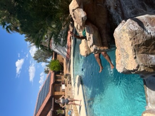 One woman stands at the edge of a pool in a bathing suit, watching another woman come down a water slide. Only the legs of the woman on the slide are visible, stretching out over the water before she lands. In the background, there's a huge teepee.